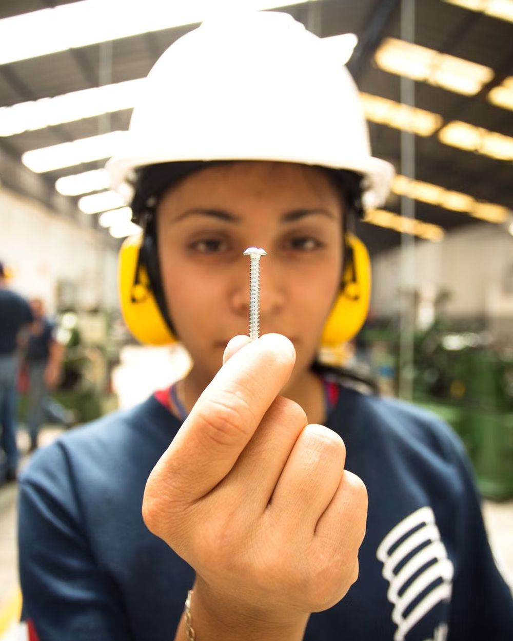 Trabajadora con casco y protección auditiva revisando una pieza en una planta industrial